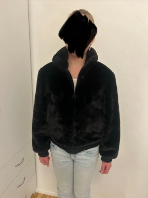 Svart fuskpäls jacka från H&M - Sammetslen lätt vadderad svart lurvig jacka från H&M med hög krage och dragkedja framtill. Jackan har ribbade muddar vid ärmslut och midja, vilket ger en schysst passform. Perfekt för dig som gillar en chill och cozy stil under kyliga dagar. Skriv om ni har någon fråga ❤️
