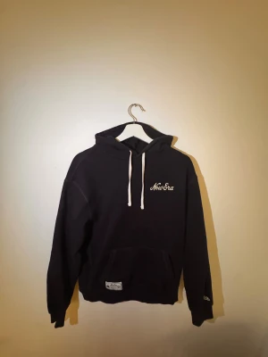 Mörkblå  hoodie från New Era med huva - Mörkblå hoodie från New Era i storlek S med vit snörning i huvan och broderad logga på bröstet. Tröjan har en klassisk känguruficka framtill och mjukt fleecefoder på insidan. 