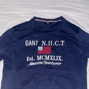 Marinblå stickad tröja från GANT - Snygg marinblå stickad tröja från GANT med broderad amerikansk flagga och vit text på bröstet. Tröjan har rund hals, ribbade muddar och klassisk sportig vibe. Perfekt för dig som gillar preppy stil och vill ha något som sticker ut.