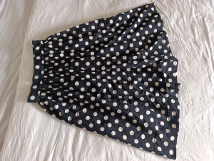 Svart prickig kjol från H&M - Snygg svart medellång kjol från H&M med vita prickar. Kjolen har en lätt utställd form och är gjord i 100% bomull, vilket gör den både luftig och bekväm. Perfekt för dig som gillar retrovibbar och vill sticka ut med ett klassiskt mönster.