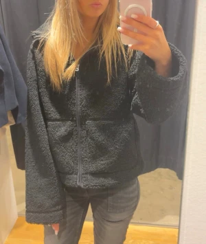 Svart teddyjacka med dragkedja - Mysig svart teddyjacka med breda ärmar och dragkedja framtill. Jackan har en oversized passform och två stora fickor på framsidan. Perfekt för att hålla sig varm och ändå se chill ut.