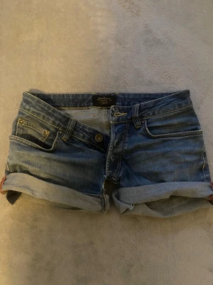 Blå lowwaisted jeansshorts  - Snygga blå jeansshorts från Premium Jeans med klassisk femficksdesign och uppvikta benslut. Shortsen har gul kontrastsöm och metallknappar framtill. Perfekta för dig som gillar en chill och avslappnad stil under varma dagar. (klippt själv)