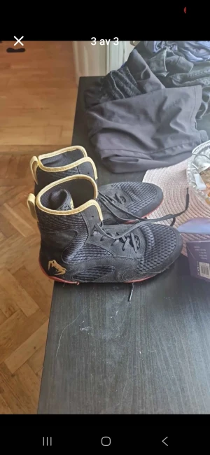 Svarta höga träningsskor med gul kant - Säljer ett par svarta höga träningsskor med gul kant och snörning. Skorna har meshpaneler för extra ventilation och en röd detalj på sulan. Perfekta för dig som vill ha en sportig look och bra stöd runt ankeln.boxningskor storlek 41.5 köpte från jabb för 1000