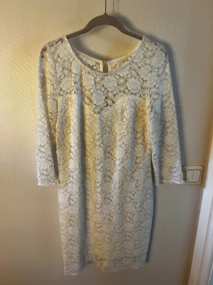 Vit spetsklänning med dragkedja - Säljer en vit kortklänning i spets med blommigt mönster och trekvartsärm. Klänningen har rund halsringning och en snygg dragkedja med guldfärgade detaljer baktill. Använd 1 gång, ev lite stor i storlek 