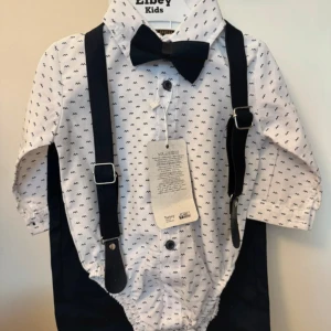Libey Kids kostymset med hängslen 68 - Supersött kostymset från Libey Kids med vit skjorta med små svarta mönster, svarta byxor, matchande svarta hängslen och fluga. Skjortan har knappar framtill och klassisk krage. Perfekt för små stiliga tillfällen! Helt nytt oanvänt 