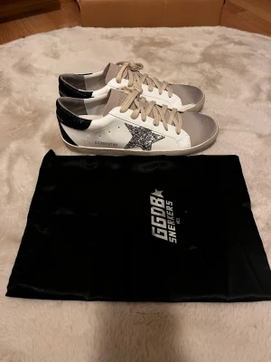 Golden Goose Superstar sneakers glitter - Golden Goose Superstar sneakers i vitt skinn med grå mockadetaljer på tån och svarta glittriga partier på hälen. Sidan har en silverglittrig stjärna och beige snörning. Klassisk låg modell med platt sula och rund tå. Snygg GGDB-påse medföljer.                           Säljer då dom är för stora 