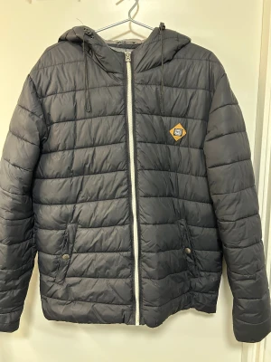 Svart pufferjacka från Only & Sons - Svart pufferjacka med huva från Only & Sons. Jackan har quiltad design, dragkedja framtill och två fickor med knappstängning. Perfekt för kyliga dagar och har en stilren look med diskret logga på bröstet.