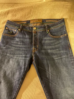 Jacob Cohën blå jeans med gröna detaljer - Snygga blå jeans från Jacob Cohën med gröna sömmar och broderier, samt en unik grön patch med logga bak. Klassisk femficksmodell med orange kontrastsömmar och rak passform. Jeansen är tillverkade i mjukt denimtyg och har metallknapp framtill. Nästan helt nya, passar mig som är 185 lite baggy men snyggt 