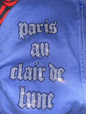 Bling lucid tjock tröja i nästan nytt skick  - Blå bling lucid tjocktröja  med dragkedja och huva, dekorerad med strass som bildar texten 'paris au clair de lune' och motiv av Eiffeltornet och månen. Hoodien har en cool vibe och sticker ut med sina glittriga detaljer. Perfekt för dig som gillar statement-plagg och Paris-inspirerad stil.