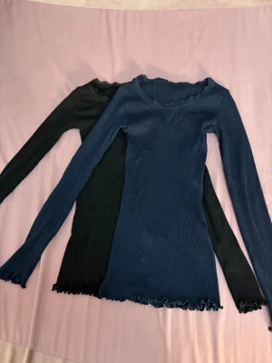 Två långärmade toppar från Rosemunde - 125kr för båda men 55kr per en! Säljer två snygga långärmade toppar från Rosemunde, en i marinblått och en i svart. Båda har ribbad struktur, spetsdetaljer vid halsringningen och vågig kant vid ärmslut och nederkant. Materialet är en lyxig mix av silke och bomull som känns skönt mot huden.