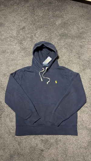 Marinblå hoodie från Polo Ralph Lauren - Snygg marinblå hoodie från Polo Ralph Lauren med klassisk gul broderad logga på bröstet. Tröjan är i storlek S men passar xs, den är i väldigt bra skick. Hör av er vid frågor ✅ny pris 1499kr