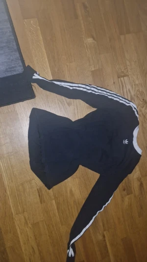 Svart långärmad Adidas tröja - Svart långärmad tröja från Adidas med klassiska vita ränder längs ärmarna och vit krage. Tröjan har Adidas logga på bröstet och är gjord i mjuk bomull. Perfekt för dig som gillar sportig och clean stil.Den är för stor för mig och bara används några gånger