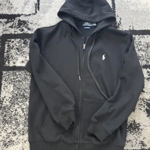 Polo Ralph Lauren hoodie - Svart hoodie från Polo Ralph Lauren med dragkedja och huva. Klassisk vit logga på bröstet och ribbade muddar. Tröjan har känguruficka och är i bomullsmaterial, perfekt för en chill och stilren look. Storleken är M men jag skulle säga att den är S