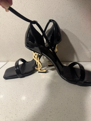Svarta Saint Laurent sandaler med YSL-klack - Säljer ett par svarta sandaler från Saint Laurent med ikonisk YSL-klack i guld. Skorna har smala remmar, fyrkantig tå och hög klack. Tillverkade i blankt skinn som ger en lyxig vibe. Perfekta för dig som vill sticka ut med designerstil.