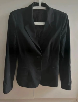 Svart kavaj från Mexx - Snygg svart kavaj från Mexx med klassisk passform och två knappar framtill. Fodret är i blankt material och kavajen har två fickor fram. Diskret broderad Mexx-logga på ärmen. Perfekt för dig som gillar stilrena plagg