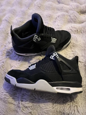 Svarta Nike Air Jordan 4 sneakers - Nike Air Jordan 4 sneakers i svart med vita och grå detaljer. Klassisk meshpanel på sidorna, snörning och ikonisk Jumpman-logga på plösen. Snygg kontrast mellan svart mocka och textil, samt vit och grå platt sula. Perfekt för dig som gillar streetwear och basketstil.