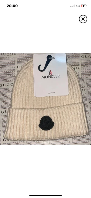 Vit ribbad Moncler mössa - Snygg vit ribbstickad mössa från Moncler med uppvikt kant och svart logopatch framtill. Perfekt för dig som vill ha en clean och lyxig look. Materialet är mjukt och värmande ull, och mössan har en klassisk form som passar till allt.