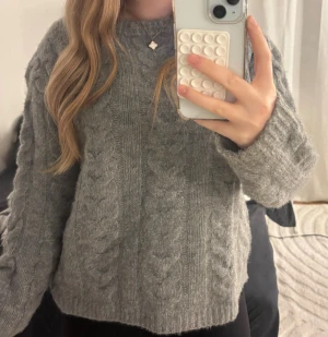 Grå stickad tröja från Lager 157 - Mysig grå stickad tröja med kabelstickat mönster framtill och på ärmarna. Tröjan har en rund hals och långa ärmar, perfekt för kyliga dagar. Den har en loose passform och är tillverkad i ett mjukt material som känns skönt mot huden.