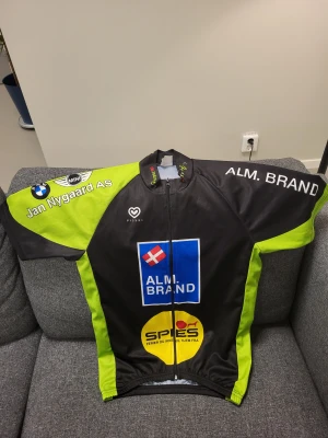Svart och neongul cykeltröja Pissei M - Cykeltröja från Pissei i svart och neongul med tryckta loggor och sponsorer. Tröjan har korta ärmar, hel dragkedja fram och praktiska bakfickor. Materialet är lätt polyester som andas, perfekt för träning och cykling. Snygg design med kontrastpaneler och sportig look.