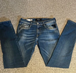 Replay Anbass Hyperflex jeans blå - Snygga Replay Anbass Hyperflex jeans i klassisk blå tvätt med coola slitningar och kontrastsömmar. Modellen har fem fickor, normal passform och stretchigt material som ger grym komfort. Perfekt för dig som gillar stilrena och moderna jeans.