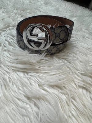 Guci bälte  - Säljer ett snyggt bälte med klassiskt monogrammö i beige och blått. Bältet har ett stort, rundat spänne i blank silverfärg. Materialet är skinn med canvas-ytan och bältet har en stilren, modern look.