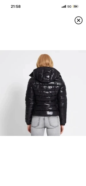 Glansig jacka - Säljer en svart pufferjacka med glansig finish och stor huva. Jackan har quiltade sektioner och är långärmad, perfekt för kalla dagar. Den har en modern, kortare passform som ger en snygg siluett och funkar till jeans eller andra byxor.