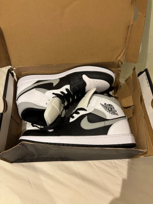 Nike Air Jordan 1 svart/vit/grå 43.5 - Nike Air Jordan 1 sneakers i svart, vit och grått med klassisk high-top siluett. Skorna har snörning, perforerad tå och ikoniska Air Jordan-loggan på sidan. Swoosh i grått och mjuk vadderad plös. Kommer med originalkartong med gul Jumpman-logo. Helt nya samt äkta från Nike hemsida! Säljes då storleken inte passade mig