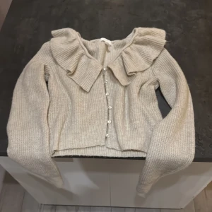 Beige stickad kofta med volang från H&M - Superfin beige stickad kofta från H&M med stora volanger längs kragen och knäppning framtill. Koftan har långärmad passform och är ribbstickad för en mysig och trendig look. Perfekt att slänga på sig när det är lite kyligare ute.💞💞