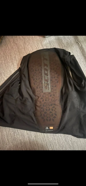 Svart skyddsväst från Scott M/L - Skyddsväst från Scott i svart med meshpaneler och integrerat ryggskydd. Västen har ett sportigt snitt och är tillverkad i syntetmaterial för att ge bra ventilation och komfort vid träning eller ridning. Snyggt geometriskt mönster på ryggskyddet och diskret logga framtill.