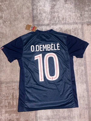 PSG Ousmane Dembélé matchtröja Nike - Paris Saint-Germain fotbollströja med O.Dembélé och nummer 10 på ryggen. Tröjan är marinblå med geometriskt mönster i rött och blått framtill, PSG-emblem på bröstet och Nike-logga. Materialet är lätt och andas, perfekt för träning eller match.