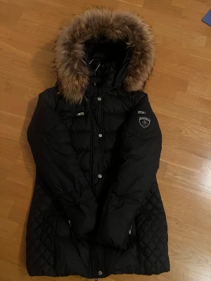 Svart Rockandblue dunjacka med päls - Snygg svart dunjacka från Rockandblue med fluffig brun päls på huvan. Jackan har quiltade detaljer, flera dragkedjor och tryckknappar framtill. Märkespatch på ärmen och lång passform som håller dig varm hela vintern.