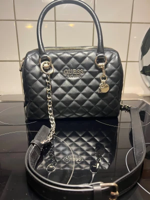 Svart quiltad handväska från Guess -  svart handväska från Guess med quiltat mönster och silverfärgade detaljer. Väskan har både handtag och avtagbar axelrem med kedja, samt dekorativt hängsmycke. Perfekt storlek för det viktigaste och stilren look.