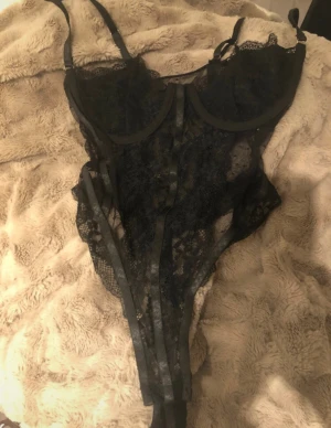 Svart spetsig korsett med justerbara band - Säljer en svart korsett/bodymed genomskinlig spets och snygga detaljer i satin. Modellen har justerbara axelband och markerade kupor för extra stöd. Spetsen har ett elegant mönster och kanten är vågig för en extra touch. Perfekt för att lyfta din outfit. ej använt då jag köpte fel storlek 