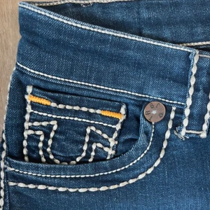 True Religion Joey jeans med kontrastsömmar - Blå True Religion Joey jeans med tydliga vita kontrastsömmar och orange detaljer vid fickorna. Klassisk femficksmodell med breda ben och ikonisk broderad logga på bakfickorna. Tjock denimkvalitet och patch med Buddha på linningen,en ersättning knapp fram. Kan tänka mig att sänka priset vid snabb affär 🥰
