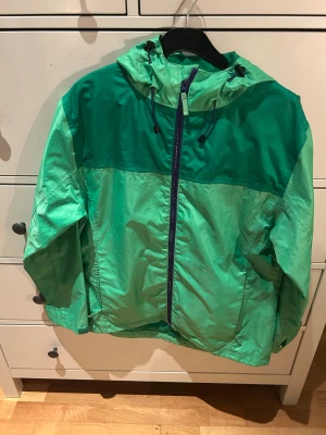 Grön vindjacka från Everest - Snygg grön vindjacka från Everest i storlek 38. Jackan har två olika gröna nyanser, huva med snörning och hel dragkedja framtill. Perfekt för blåsiga dagar och outdoor-aktiviteter. Lätt och smidig att bära med sig, med klassisk sportig look. Är liten i storleken.
