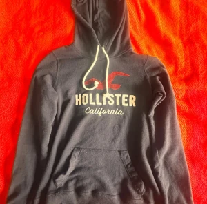 Mörkblå hoodie från Hollister S - Mörkblå hoodie från Hollister med vit snörning och tryck i vitt och rött på bröstet. Klassisk känguruficka framtill och mjukt material i bomull och polyester. Perfekt för chill dagar och har en skön huva som ger extra mysfaktor.
