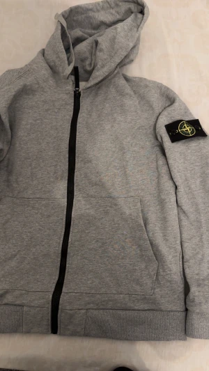 Grå hoodie med dragkedja från Stone Island  - Snygg grå hoodie från Stonesland med svart dragkedja och huva. Tröjan har en klassisk patch på vänster ärm, två fickor framtill och ribbade muddar. Perfekt för chill dagar och streetwear-stil.