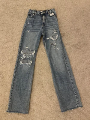 Blå ripped jeans från Zara, strl 38 - Säljer ett par blå jeans från Zara i storlek 38 med hög midja och raka ben. Jeansen har flera stora slitningar och hål framtill och fransade benslut. Jag är 170cm. 