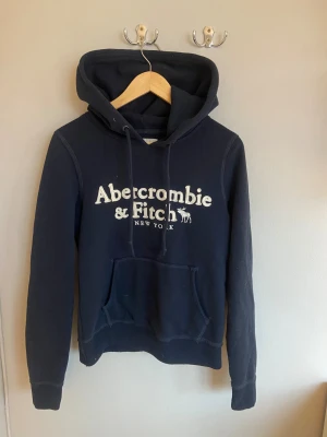 Mörkblå hoodie från Abercrombie & Fitch - Snygg mörkblå hoodie från Abercrombie & Fitch med vit broderad logga på bröstet. 