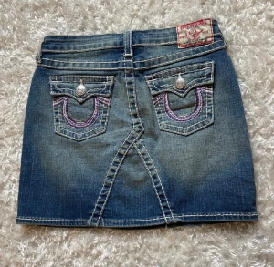 True Religion jeanskjol med kontrastsömmar - Säljer en snygg blå jeanskjol från True Religion med tydliga vita kontrastsömmar och lila broderi på bakfickorna. Kjolen har klassiska fickor fram och bak, samt en cool patch med logga bak i midjan. Perfekt för dig som gillar streetstyle och denim. Storlek 14