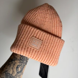 Ljusrosa Acne Studios peach mössa i 100% ull - Säljer en ribbstickad peach mössa från Acne Studios med det ikoniska ansikts-emblemet framtill. Mössan är tillverkad i 100% ull och har en bred uppvikt kant för extra stilpoäng. Perfekt för dig som vill ha en clean look med lite edge.