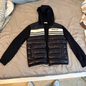 monclear cardigan - Svart pufferjacka från Moncler med huva och vita ränder över bröstet. Jackan har dragkedja framtill, två fickor och Moncler-logga på ärmen. Kroppen är quiltad och glansig medan ärmarna är i mjukare material. Perfekt för dig som vill ha en clean look