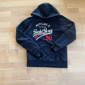 Mörkblå hoodie Jack & Jones - Snygg mörkblå hoodie från Jack & Jones med tryck i vitt och rött på bröstet. Tröjan har huva med snörning och ribbade muddar.  Materialet är mjuk bomull och polyesterblandning. Skriv vid minsta fundering