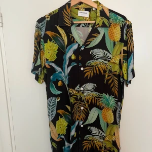 Svart tropisk skjorta från Lindbergh - Svart skjorta från Lindbergh med tropiskt mönster i grönt, gult och blått. Relaxed fit med korta ärmar och knappar framtill. Materialet är mjukt och lätt, perfekt för varma dagar. Snygg krage och detaljerad print med ananas och blad.