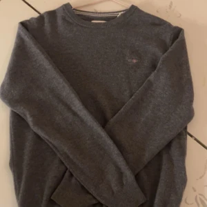 Mörkgrå stickad tröja GANT S - Mörkgrå stickad tröja från GANT i storlek S. Klassisk rund hals med ribbade muddar och diskret logga broderad på bröstet. Perfekt för dig som gillar stilrena och enkla plagg med snygg passform.