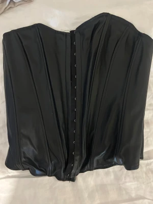 Svart korsett i skinnimitation från Zara - Säljer en svart korsett från Zara i storlek XS. Toppen har en snygg skinnimitation med markerade sömmar och hakknäppning framtill. Den är axelbandslös och har en stilren, strukturerad form som ger en edgy vibe. Perfekt för dig som vill sticka ut med en cool look.