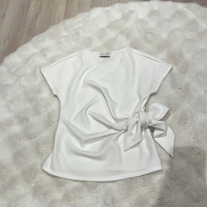 Vit omlottopp med knyt från Anna Field - Supersnygg vit topp från Anna Field med omlottdesign och bred knytning i sidan. T-shirtrmad och rund halsringning. Toppens material har en lätt glansig finish som ger en fräsch look. Perfekt att styla med jeans eller kjol för en clean vibe.