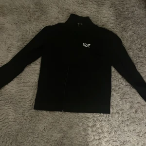 Svart zipjacka EA7 Emporio Armani - Svart zipjacka från EA7 Emporio Armani med stilren design och logga på bröstet. Jackan har hög krage, långa ärmar och dragkedja framtill. Materialet är mjukt och känns sportigt, perfekt för en chill och snygg look.
