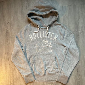 Grå hoodie från Hollister med tryck - Säljer en grå hoodie från Hollister med vit broderad text och palmtryck på bröstet. Tröjan har huva med snörning, magficka och ribbade muddar. Perfekt för chill dagar och har en mjuk insida i bomullsmix. Klassisk surf club-stil.