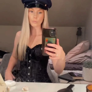 Svart korsettväst med snörning - Snygg svart väst i korsettstil med snörning framtill och dekorativa knappar. Västen har en v-ringad urringning och är figurnära med en glansig finish. Perfekt för dig som vill sticka ut och skapa en edgy look.